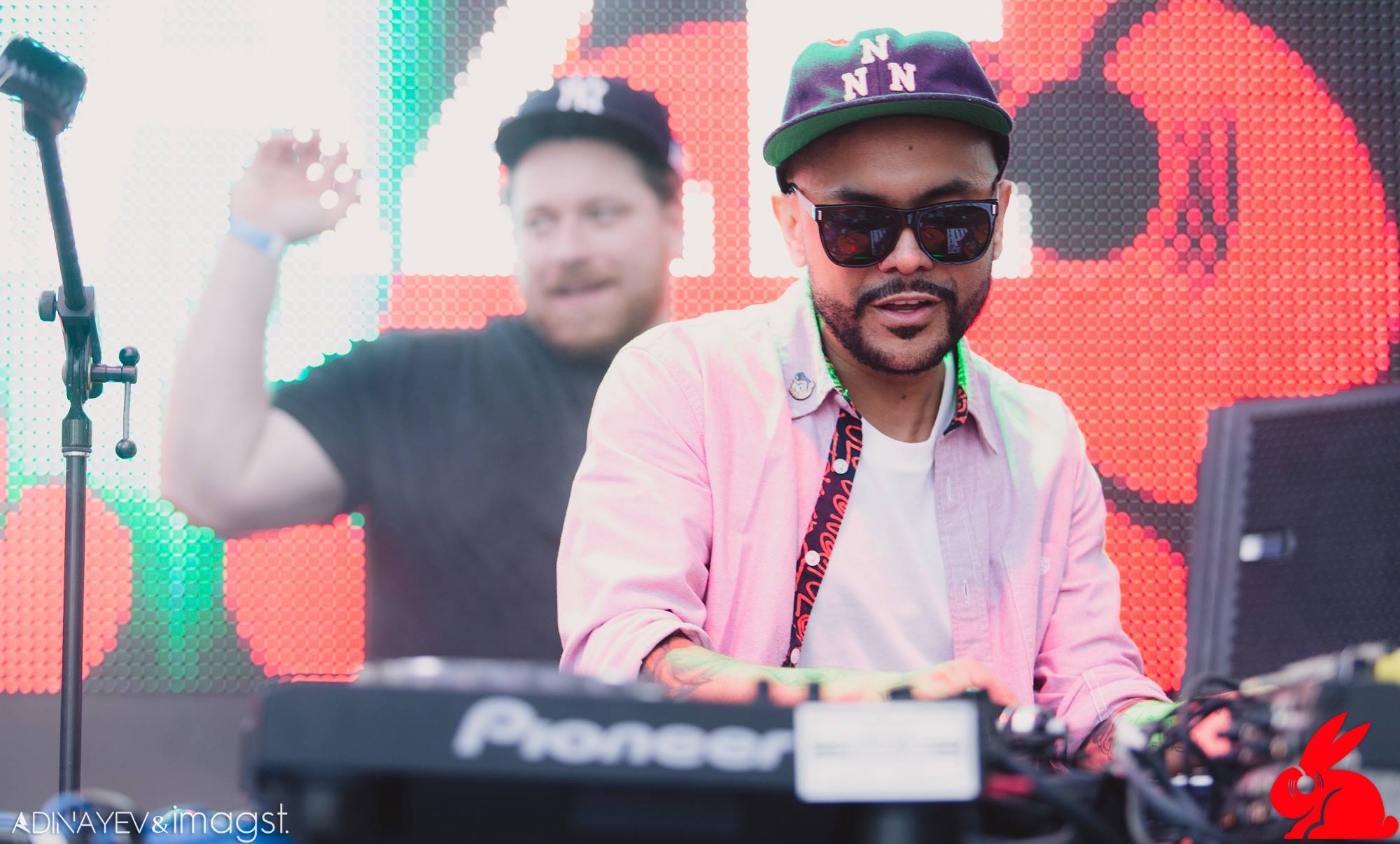 DJ in a pink shirt and cap behind the booth in front of a red LED screen