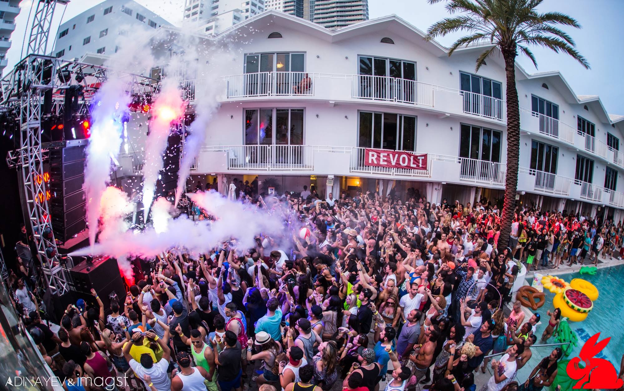 Daytime pool party crowd with confetti cannons firing under a REVOLT banner and palm trees