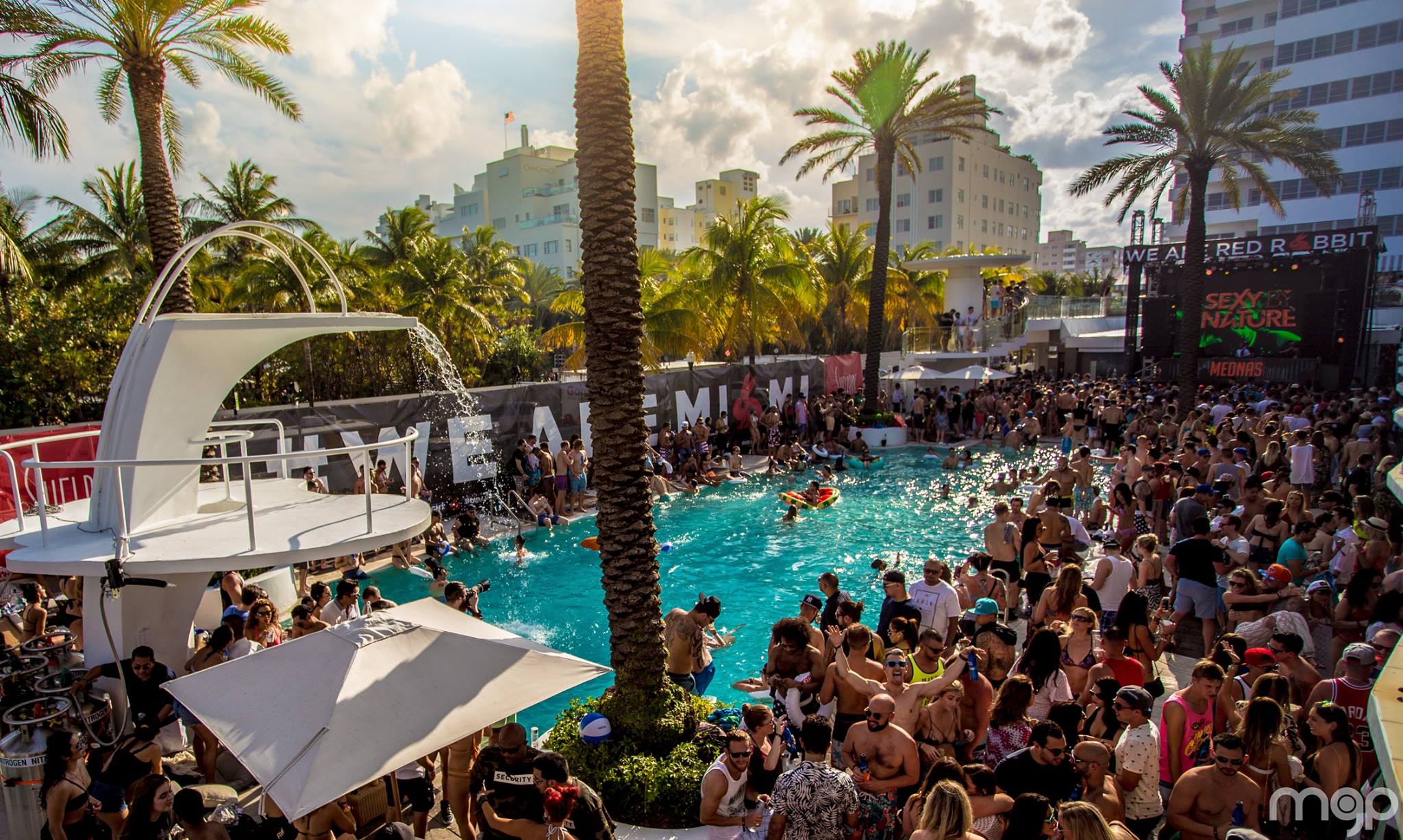 Sun-drenched pool deck with a WE ARE MIAMI banner, palm trees, and a packed daytime crowd