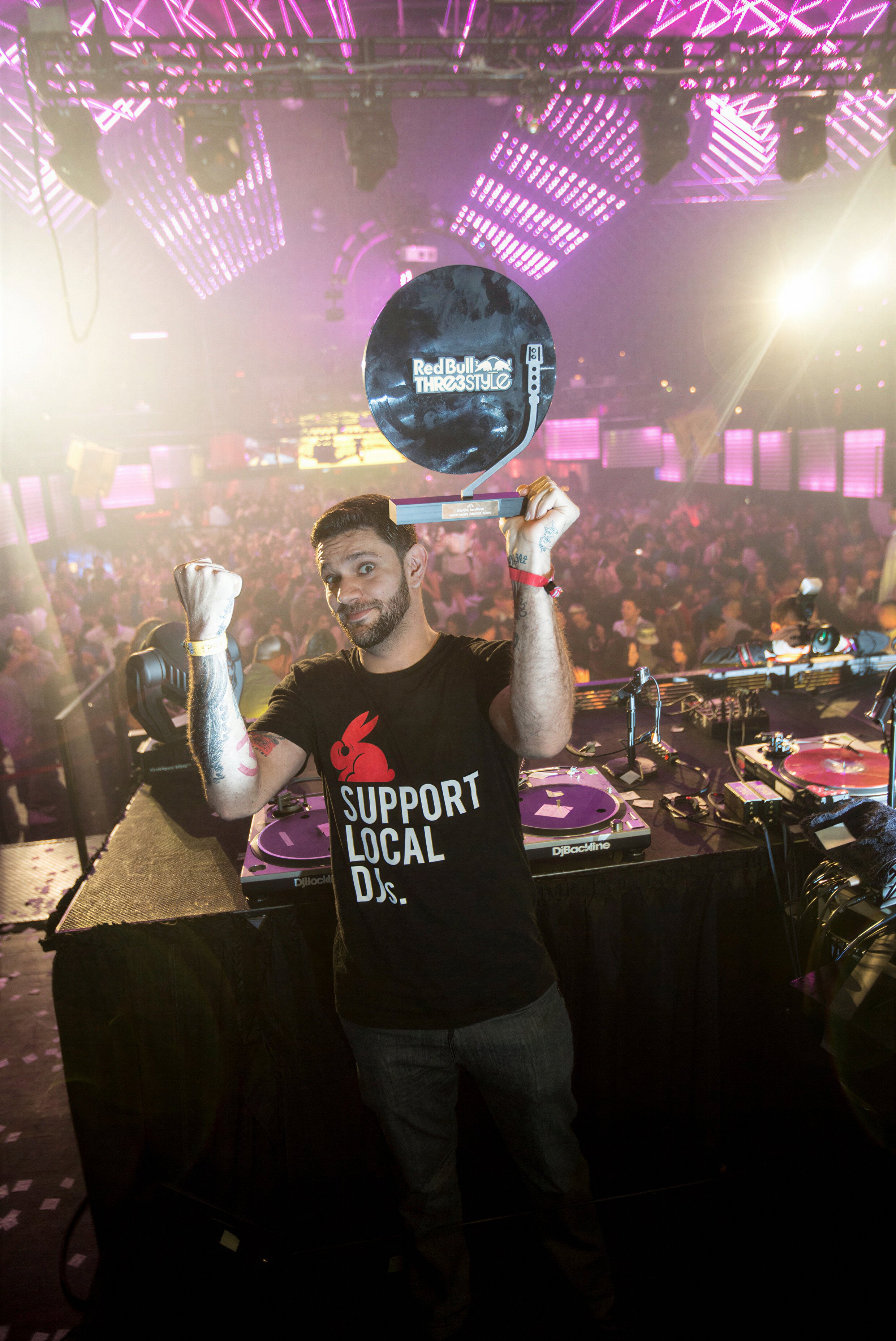 DJ in a Support Local DJ tee holding a record above his head in a crowd under purple light