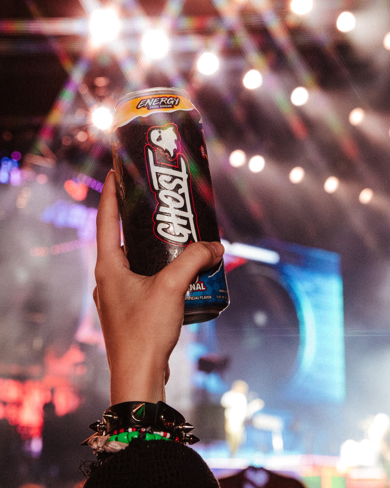 Hand raising a GHOST Energy can toward the stage lights at night